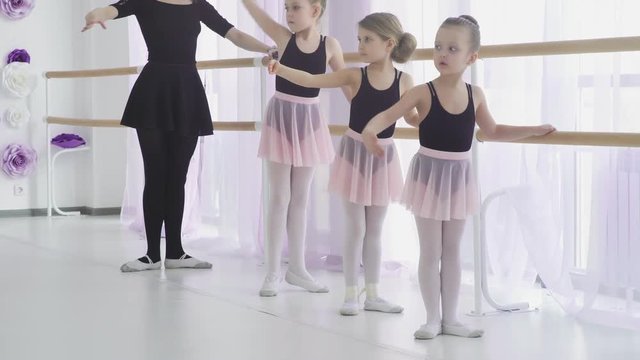 Little girls are having classical ballet lesson learning leg movements with teacher in art studio.