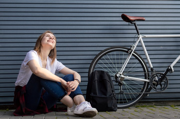 young stylish girl sits near the bike and backpack and dreams, smiles against the gray wall