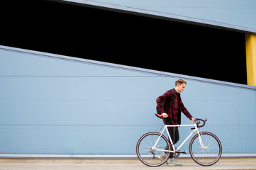 young stylish guy walks with a white bicycle in front of a blue wall, a student with a backpack...