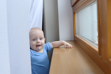 One year old baby boy with blue eyes is playing with curtains near window. Need to be careful with choosing children playground