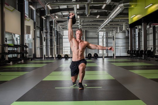Tall shirtless muscular caucasian athlete  does an overhead kettlebell lift with a lunge inside a gym 