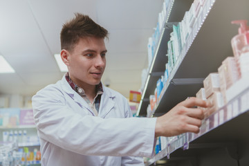 Low angle shot of an attractive male pharmacist filling shelves of drugstore with new products for sale. Male chemist working at pharmacy. Health care, medicine, consumerism concept