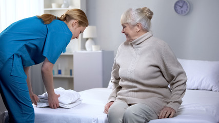 Nurse preparing clean bed-linen to elderly female patient in medical center