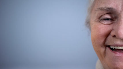Fototapeta premium Half-face of smiling old woman grey background, social security, positive mood