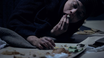 Hungry homeless person lying on ground with garbage and greedily eating orts