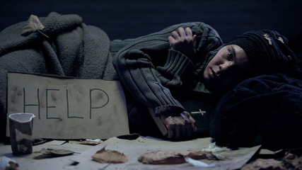 Depressed homeless person with bible and help sign on cardboard lying on floor
