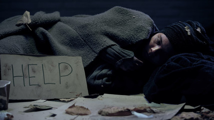 Homeless bum with help sign on board lying in dirty place, shivering with cold