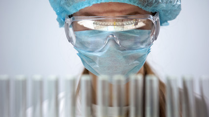 Female chemist in protective mask looking at test tubes, biological research