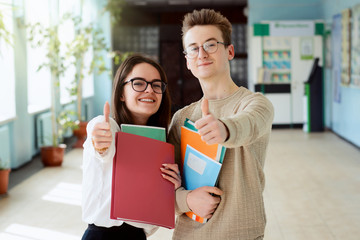 Two happy students in good mood showing thumbs up to camera standing in campus of university,...