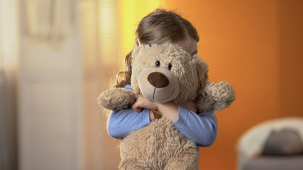 Preschool shy girl hiding behind teddy bear, childish psychological problems - obrazy, fototapety, plakaty