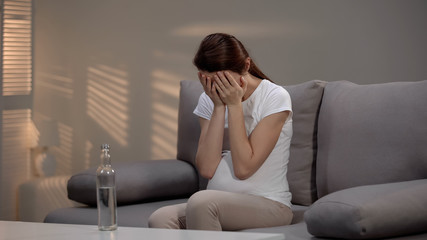 Pregnant woman crying, vodka standing on table, alcohol addiction impact on baby