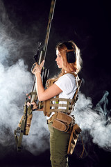 Adorable riot woman with blonde long hair dressed in military style, wearing plate carrier and warbelt on her thin waist, holding snipper rifle in hands, posing in studio over foggy dark background