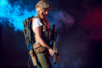 Female storm trooper in military camouflage uniform protected with helmet, body armour with submachine gun seeking aims in the smoky darkness