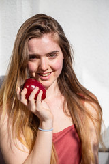 Young joyful attractive woman in casual summer dress sitting on balcony and eating tasty juicy red apple