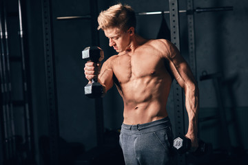 Fototapeta na wymiar Shirtless white man in sweatpants starting exercise with dumbbell weight in dark gym. Fitness motivation and muscle training concept.