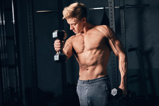 Shirtless white man in sweatpants starting exercise with dumbbell weight in dark gym. Fitness motivation and muscle training concept.
