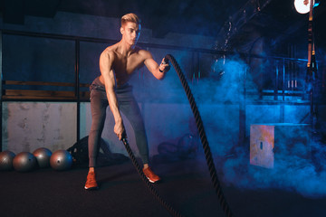 Fototapeta na wymiar Handsome muscular champion man is doing battle rope exercise while working out in dark gym, preparing to cross fit international contest