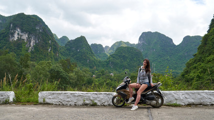 Young beautiful brunette woman sits and posing on the motorbike on the road with panoramic view to scenic landscape with green mountains. Cat Ba Island, Vietnam.