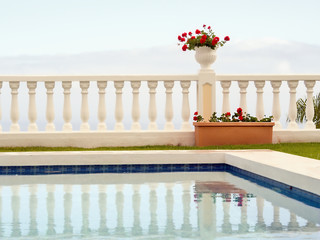 Minimalism of form and color, a terraced white space with a white pot with dark red geraniums against whitish sky.