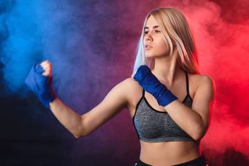 Obraz premium Attractive blonde woman fighter in boxing bandages posing in defense boxer stance isolated on dark smoky background in sport and fitness exercise workout.