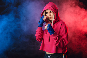 Long-haired blonde kickboxing woman in pink sweatshirt with hood on, training in fitness studio....