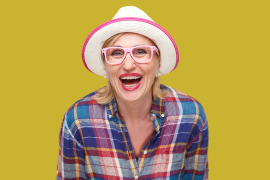 Portrait of happy modern stylish mature woman in casual style with hat and eyeglasses standing and looking at camera with surprised face and laughing. indoor studio shot isolated on yellow background.