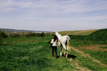 A young woman leads a white horse alongside a green field. Love to the animals. Farm and equestrian sport.