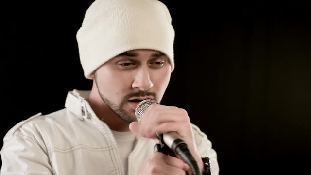 Close-up Frontman vocalist rock pop with a stylish beard in white clothes and a hat with a microphone in his hands expressively aggressively singing in the studio against the background of black walls