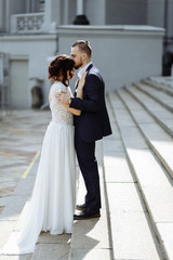 Young beautiful stylish pair of newlyweds hugs each other on stairs near building with columns