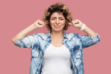 Portrait of confused young woman with curly hairstyle in casual shirt standing with fingers on her ears and looking at camera with dissatisfied face. indoor studio shot, isolated on pink background.