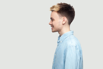 Profile side view portrait of happy handsome young man in light blue shirt standing with toothy smile and looking forward with happiness. indoor studio shot on grey background copy space.