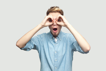 Portrait of shocked handsome young man in light blue shirt standing with hands on eyes binoculars gesture and looking at camera with surprised face. indoor studio shot on grey background copy space.