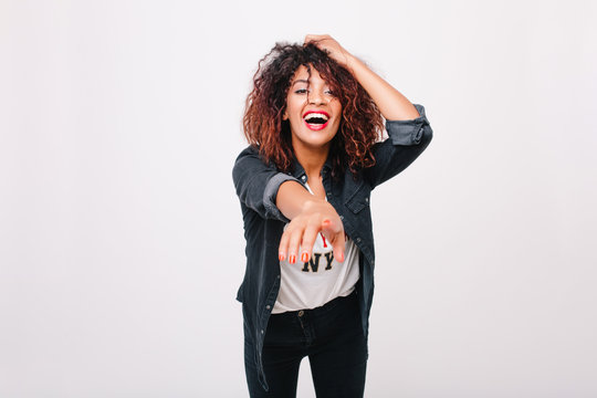 Enchanting black young woman in denim jacket pointing with finger to camera. Refined curly girl with excited smile posing in trendy attire standing in studio.