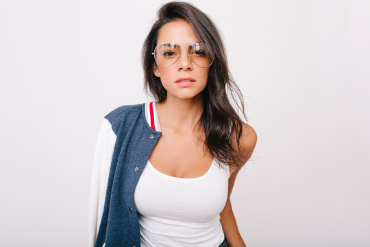 Close-up portrait of stunning dark-haired girl in white tank-top looking with interest to camera. Indoor photo of adorable european lady in stylish glasses chilling on photoshoot.