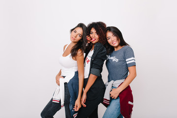 Refined girls in trendy t-shirts posing together in studio and laughing. Indoor photo of fascinating asian lady spending time with friends enjoying photoshoot on white background.