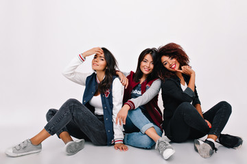 Laughing beautiful ladies in trendy sneakers sitting with legs crossed after photoshoot. Indoor portrait of asian girl resting on the floor between latin and african friends.