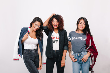 Fototapeta premium Indoor portrait of three excited female students in trendy clothes having fun together after lessons. Curly girl in denim attire spending time with brunette friends and laughing.