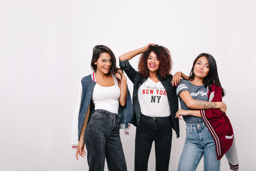 Blissful latin girl in jeans having fun with university mates posing in studio with smile. Indoor portrait of lovely african lady standing between friends and playing with curly hair.