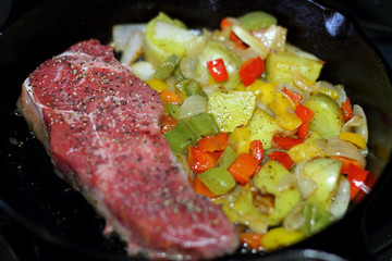 New York strip steak frying in a cast iron pan with potatoes,assorted bell peppers, and onions on the stove top.