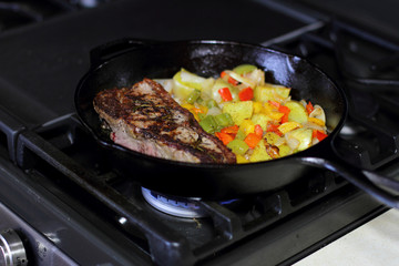 New York strip steak frying in a cast iron pan with potatoes,assorted bell peppers, and onions on the stove top.