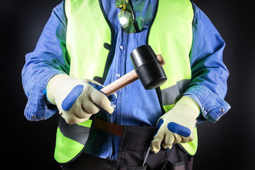 Worker in jeans shirt and green jacket holding rubber mallet.