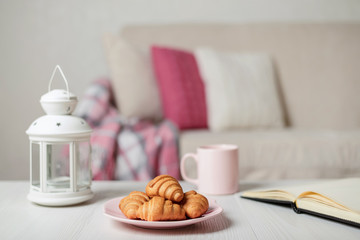 Beige sofa with plaid, colorful pillows (pink, grey, white) and coffee with croissantsin the living room.