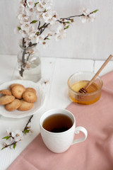 A cup of black tea on a pink napkin, homemade cookies, a bowl of honey and a glass vase of apricot bloom on the white background.