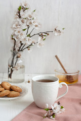 A cup of black tea on a pink napkin, homemade cookies, a bowl of honey and a glass vase of apricot bloom on the white background.