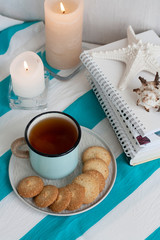 A hot mug of black tea with homemade cookies on a striped tablecloth, wax candles, decorative starfish, seashell, pile of notebooks on a white background.