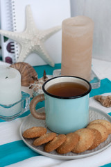 A hot mug of black tea with homemade cookies on a striped tablecloth, wax candles, decorative starfish, seashell, pile of notebooks on a white background.