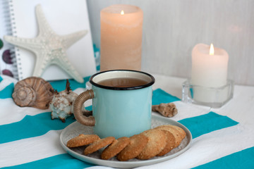 A hot mug of black tea with homemade cookies on a striped tablecloth, wax candles, decorative starfish, seashell, pile of notebooks on a white background.