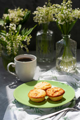 A tasty breaksfast. A plate of freshly-baked muffins with a hot cup of coffee on a white tablecloth, bouquets of lilies of the valley in glass vases on the gray background.