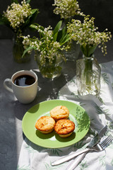 A tasty breaksfast. A plate of freshly-baked muffins with a hot cup of coffee on a white tablecloth, bouquets of lilies of the valley in glass vases on the gray background.