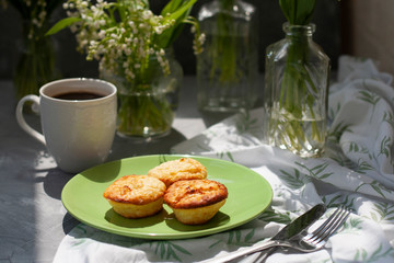 A tasty breaksfast. A plate of freshly-baked muffins with a hot cup of coffee on a white tablecloth, bouquets of lilies of the valley in glass vases on the gray background.